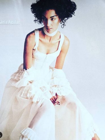 Vania Aguiar in a stylish white dress with layered ruffles and curly hair, seated against a neutral backdrop.