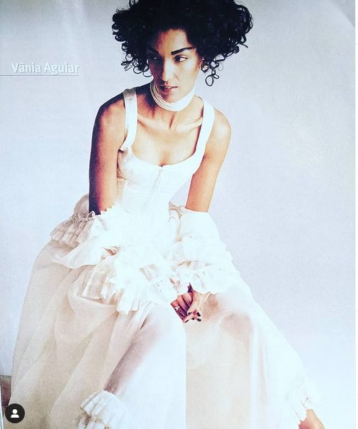 Vania Aguiar in a stylish white dress with layered ruffles and curly hair, seated against a neutral backdrop.