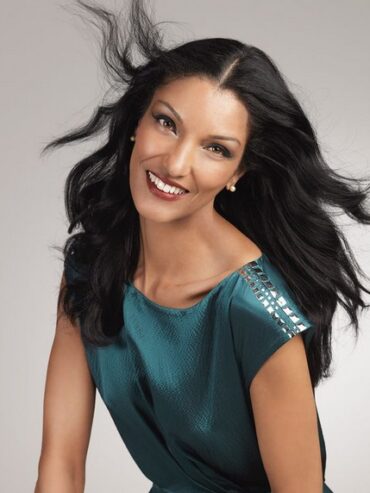 Fashion portrait of a woman with flowing black hair, wearing a teal top, smiling confidently.