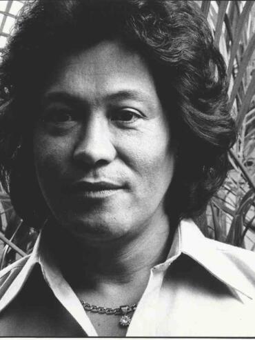 Black-and-white portrait of a woman in a collared shirt, standing against lush plant foliage.
