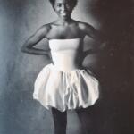 Black-and-white image of a woman wearing a strapless top and puffed skirt, posing confidently with her hands on her hips.