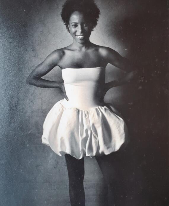 Black-and-white image of a woman wearing a strapless top and puffed skirt, posing confidently with her hands on her hips.
