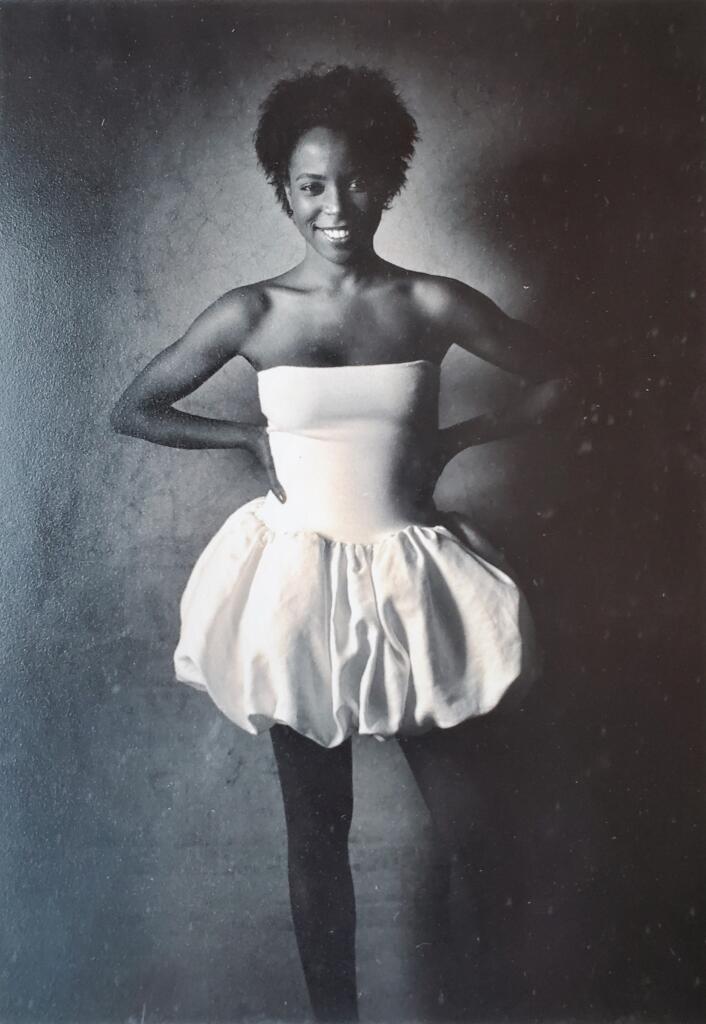 Black-and-white image of a woman wearing a strapless top and puffed skirt, posing confidently with her hands on her hips.