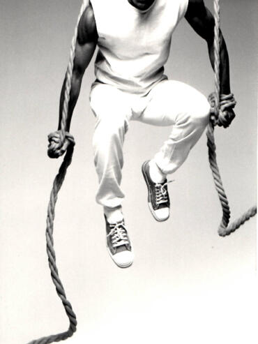 A man with an energetic expression, wearing casual clothes and sneakers, appears to be mid-air while holding onto a thick rope.