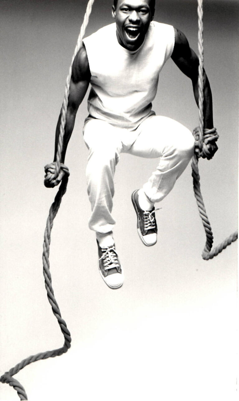 A man with an energetic expression, wearing casual clothes and sneakers, appears to be mid-air while holding onto a thick rope.