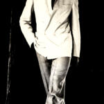 Model walking the runway in a tailored suit with a jacket draped over one shoulder in a high-fashion setting.