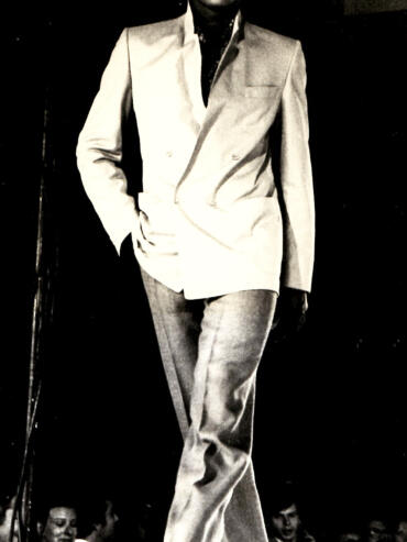 Model walking the runway in a tailored suit with a jacket draped over one shoulder in a high-fashion setting.