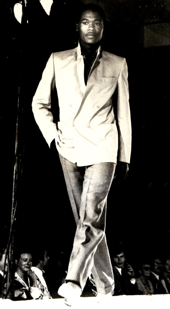 Model walking the runway in a tailored suit with a jacket draped over one shoulder in a high-fashion setting.