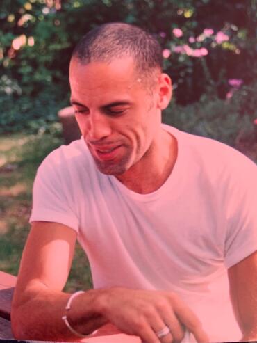 Man with closely shaved hair, smiling outdoors in a pink-tinged light.