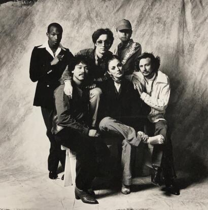 Group portrait of seven diverse individuals dressed in vintage and casual styles, posed against a textured backdrop.