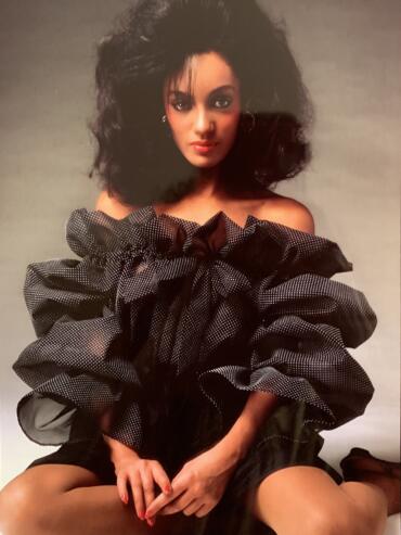 Woman with voluminous curly hair wearing an off-shoulder ruffled polka dot dress, seated with legs apart.