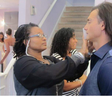 A makeup artist applies makeup to a man at an indoor event.