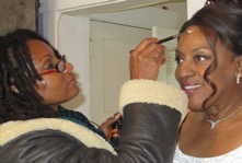 A makeup artist applying eyeliner to a smiling woman with braided hair