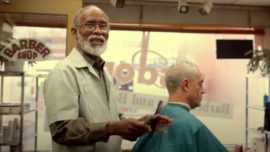 Barbershop scene with an older barber holding a hair clipper, cutting a customer's hair.