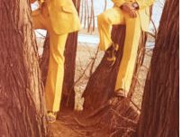 Two women in yellow suits and wide-brimmed hats sit among trees in an outdoor setting.