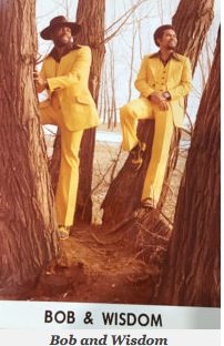 Two women in yellow suits and wide-brimmed hats sit among trees in an outdoor setting.