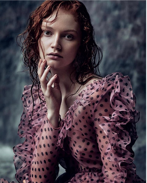 Young woman with wet red hair wearing a sheer polka dot blouse against a dark background.