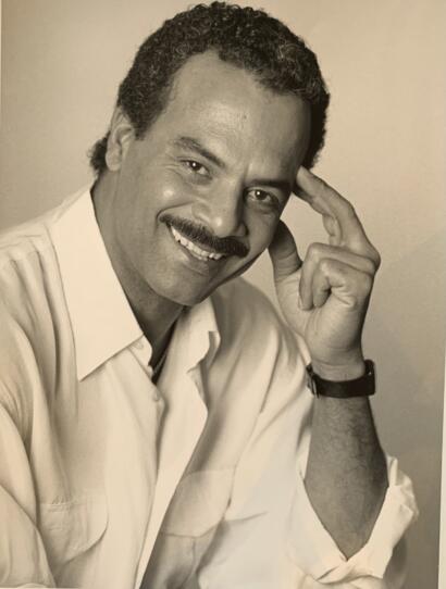 Man with curly hair, mustache, and smiling, wearing a light shirt and watch.