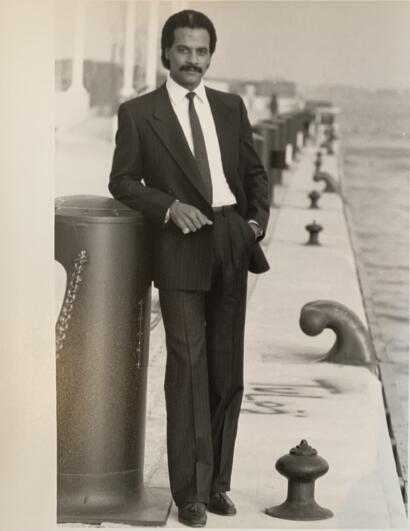 Man in a tailored suit leaning on a railing by the water, classic black-and-white photography.