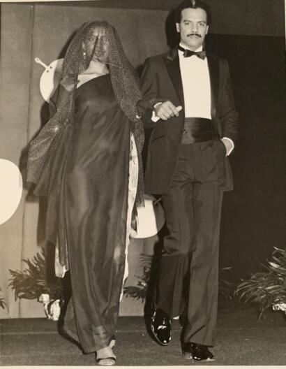 Elegant man in a tuxedo walking arm-in-arm with a woman in a flowing gown and veil.