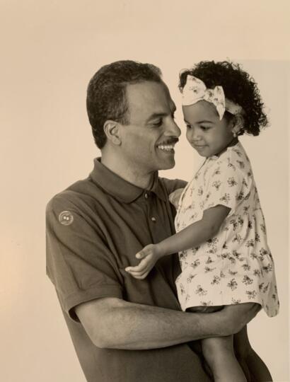 A man holding a young girl, both smiling softly, in a warm, candid moment.