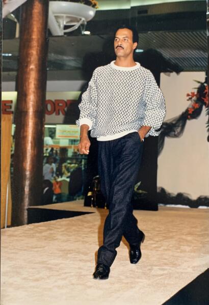 Male runway model in a patterned sweater and dark trousers walking on a fashion show stage.
