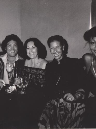 Group of five elegant women smiling at a social gathering, dressed in vintage evening attire.