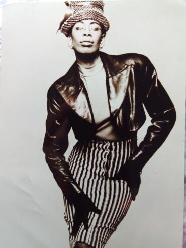 Black and white portrait of a woman in vintage fashion, wearing a hat, leather jacket, striped skirt, and earrings.