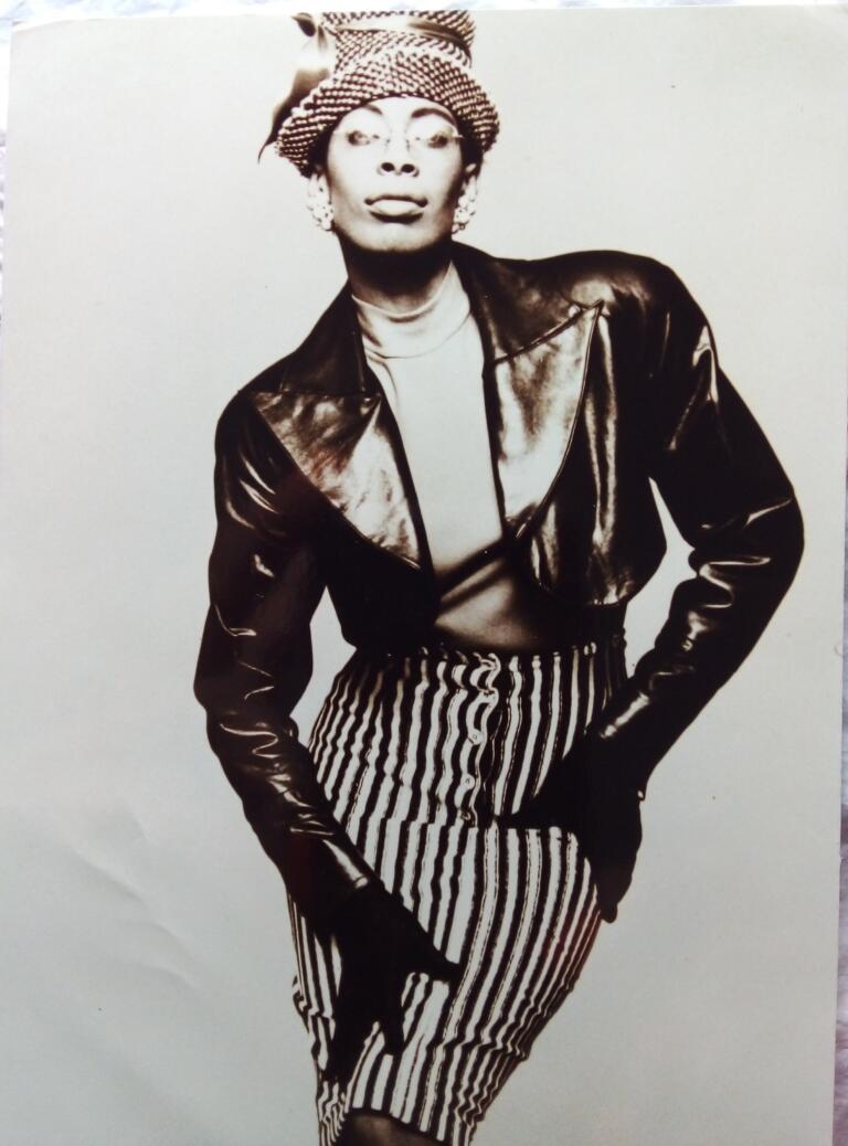 Black and white portrait of a woman in vintage fashion, wearing a hat, leather jacket, striped skirt, and earrings.