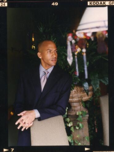 Man in a dark pinstripe suit and light-colored tie seated indoors near lush greenery.