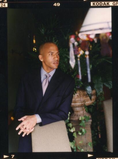 Man in a dark pinstripe suit and light-colored tie seated indoors near lush greenery.