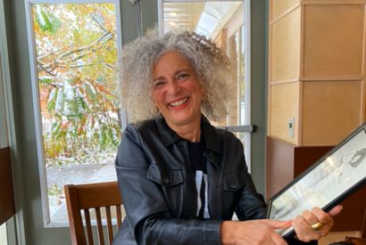 Woman with curly gray hair smiling while holding a tablet indoors.