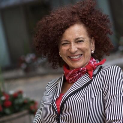 Woman with curly red hair, striped blazer, red bandana, and pearl earrings smiling.
