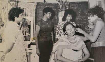 Women in a vintage hair salon, one getting a haircut, others observing and discussing.