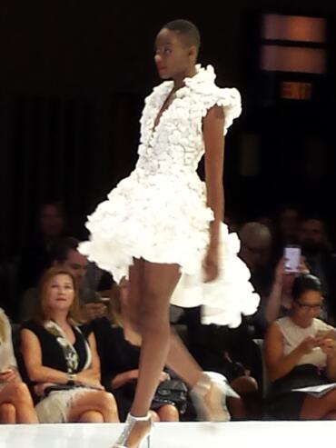 Model walks the runway in a white floral dress at a fashion show.