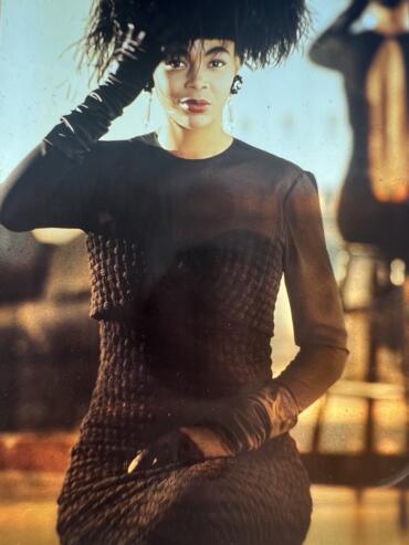 A woman dressed in elegant black attire and feathered headpiece, posing outdoors at sunset.