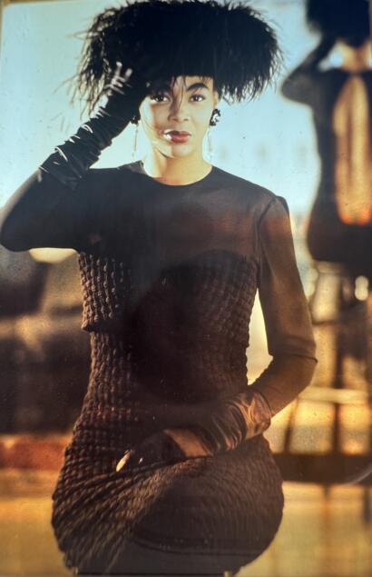 A woman dressed in elegant black attire and feathered headpiece, posing outdoors at sunset.