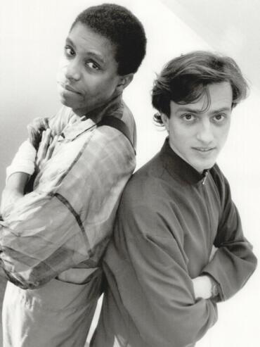 Black-and-white photo of two men standing back-to-back with arms crossed, showcasing casual fashion styles.