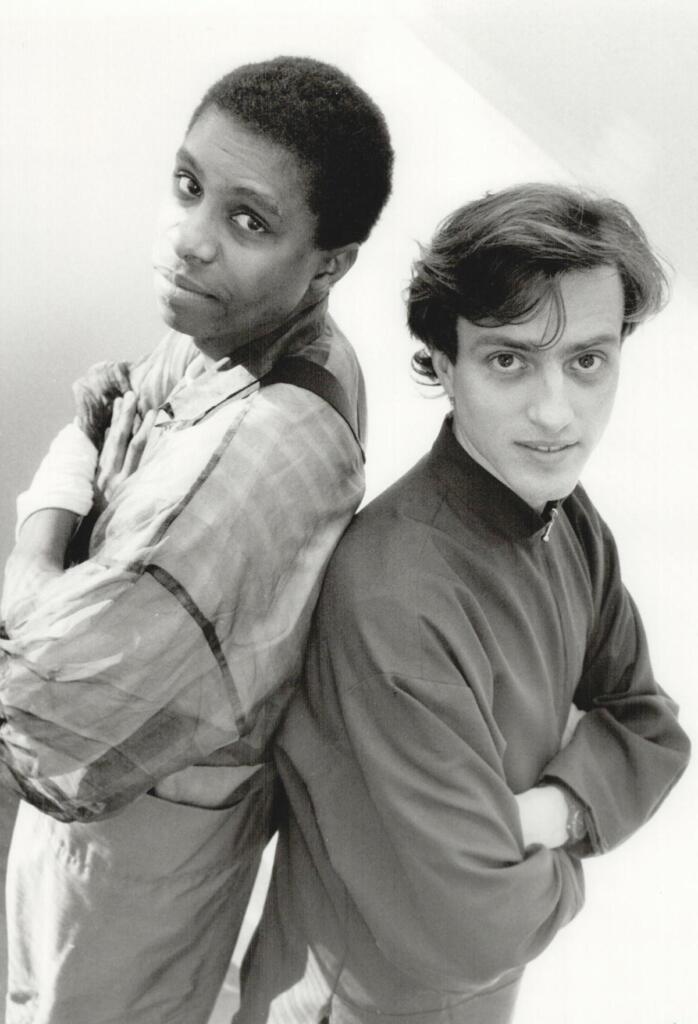 Black-and-white photo of two men standing back-to-back with arms crossed, showcasing casual fashion styles.