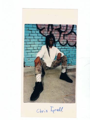 Individual with dreadlocks wearing sunglasses, patterned pants, white shirt, and polka-dot socks posing in front of graffiti wall.