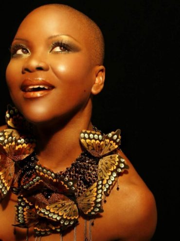 A woman with a shaved head wears butterfly jewelry, striking a thoughtful pose against a black background.