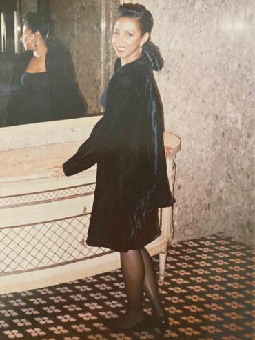 Elegant woman in a black velvet coat, standing beside a vintage dresser, smiling.