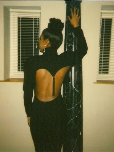 Woman in a black gown with an open back, standing beside a marble column in front of two windows.