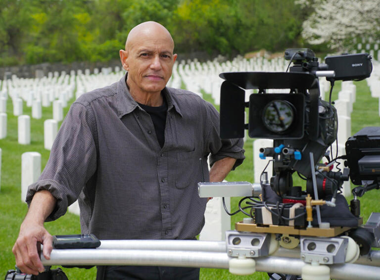 A filmmaker reviews a camera setup at a cemetery, with white headstones and greenery in the background.