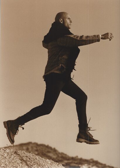 A person mid-jump on a rocky terrain, dressed in dark pants, rugged boots, and a textured jacket.