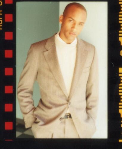 A man in a beige suit and turtleneck, posing confidently against a minimalist background.