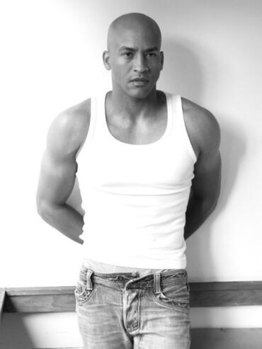 Black-and-white portrait of a person in a sleeveless top and jeans, standing with arms behind their back.