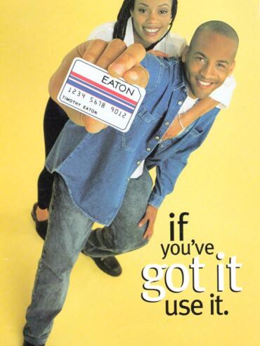 Young man holding a store card, smiling, with a woman behind him, yellow background, promotional message.