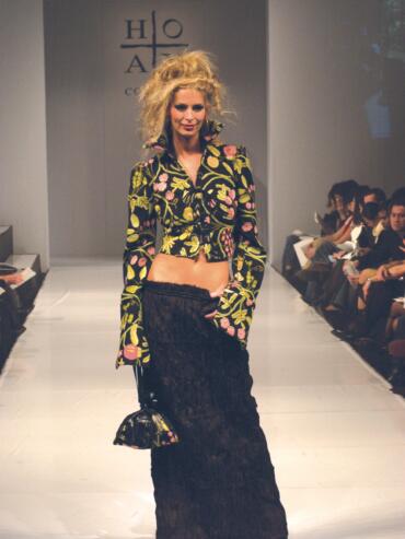 Model walking down the runway in a floral embroidered jacket with a black skirt.