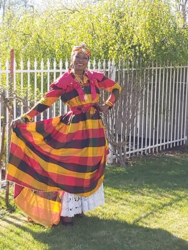 Woman standing outdoors in a colorful, striped dress with a white lace underskirt, smiling.